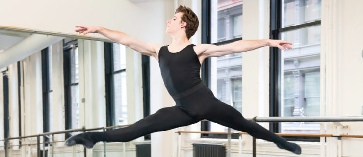 Jake Roxander, wearing black tights, ballet slippers and a black tank top, leaps with his right leg in front in a large dance studio.