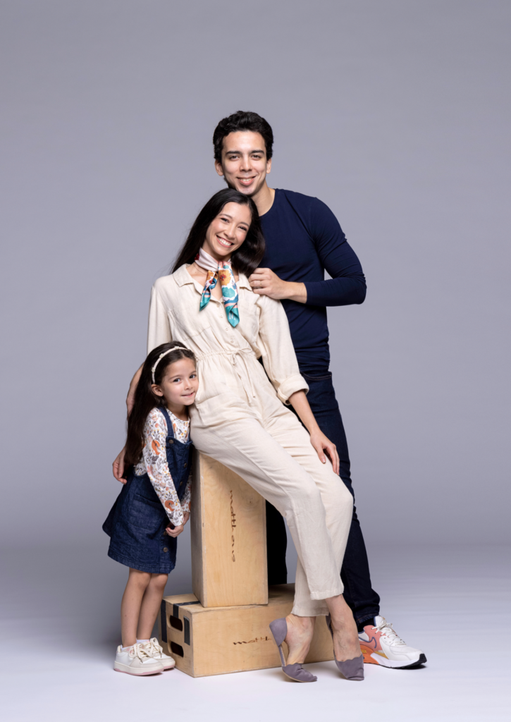 Macarena Giménez, wearing a khaki jumpsuit and multi-colored neckscarf, sits on an apple box and leans her head against Maximiliano Iglesias's right shoulder. He stands behind her and rests his head on top of her head, and wears a blue shirt, black pants and sneakers. On Gimenez's right side, her five-year-old daughter snuggles up close and rests her head on her mother's hip. She wears a blue dress and sneakers. They all look toward the camera and smile warmly.
