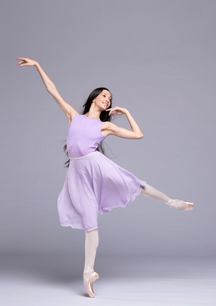 Macarena Giménez, wearing a purple dance dress, pink tights and pointe shoes, poses in a low arabesque on pointe with her left leg up. She looks over her left shoulder, raises her right arm up and pulls her left arm in, brushing her chin with her left hand.