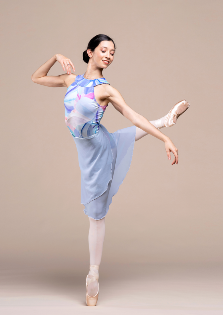 Macarena Giménez poses in an attitude derriere croisé on pointe with her right leg in back. She bends her right arm up and touches her right hand to her shoulder and extends her left arm out to the side. She wears a blue, white and pink multi-colored leotard and long gray skirt, pink tights and pointe shoes.
