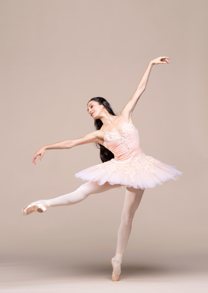 Macarena Giménez wears a pink tutu, pink tights and pointe shoes and poses on pointe in a degagé devant in effacé with her right leg lifted. She reaches her right arm out and twists her upper body slightly to the left, looking over her hand, and raises her left arm up.