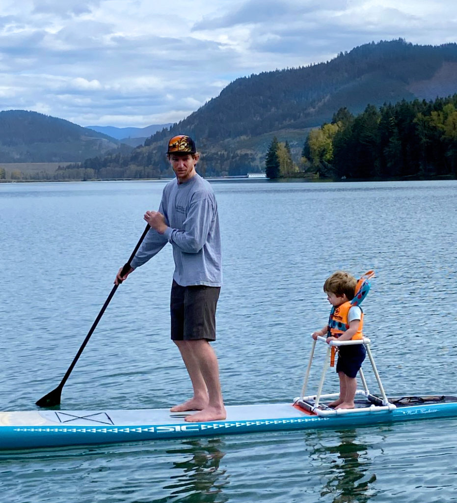A man stands on a paddleboard on a lake. Behind him is his son in a secure stand and life jacket.