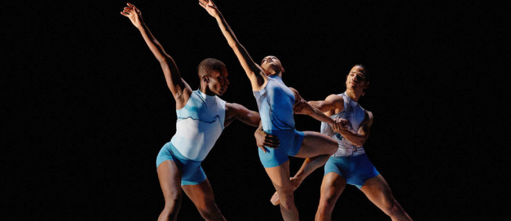 A trio of dancers—two men and one woman— perform onstage. They are dressed in blue leotards and shorts and the woman wears brown pointe shoes. The two men stand on either side of her lunging on their right leg and holding on to her waist and left arm as she does a retiré on pointe with her left foot behind her knee. She reaches her right arm high and looks toward the sky.