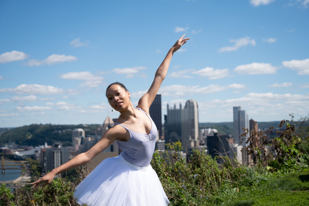 A Black ballerina outdoors in a grassy area overlooking downtown Pittsburgh.