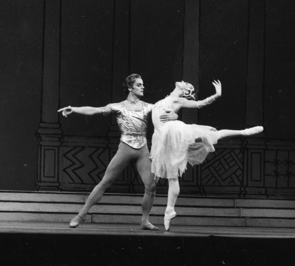 In this black and white photo, Mikhail Baryshnikov and Lesley Collier perform a pas de deux onstage in costume. Baryshnikov lunges slightly to his left and holds on to Collier's waist as she arches her back in a high arabesque. He wears a short-sleeved tunic, tights and ballet slippers, while she wears a light-colored, knee-length tutu, tights and pointe shoes.