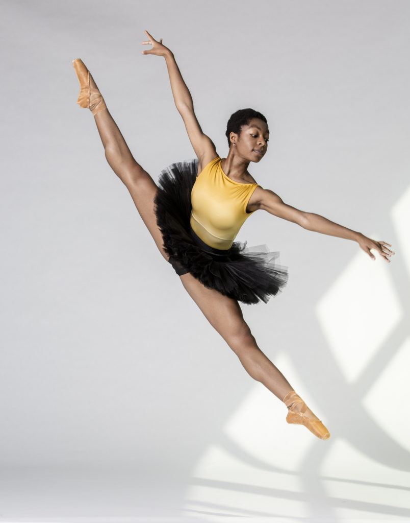Ryan Smallwood, wearing a yellow leotard, black practice tutu and tan pointe shoes, jumps and splits her legs in an ecarté derrie