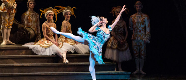 Tina Pereira performs in The Sleeping Beauty onstage. She wears a bright blue tutu and feathered headpiece, pink tights and pink pointe shoes. She performs a first arabesque with her right leg behind her, and she bends her right arm in so that her hand grazes her ear. Behind her, several dancers in grand costumes sit and stand on a staircase and watch her.