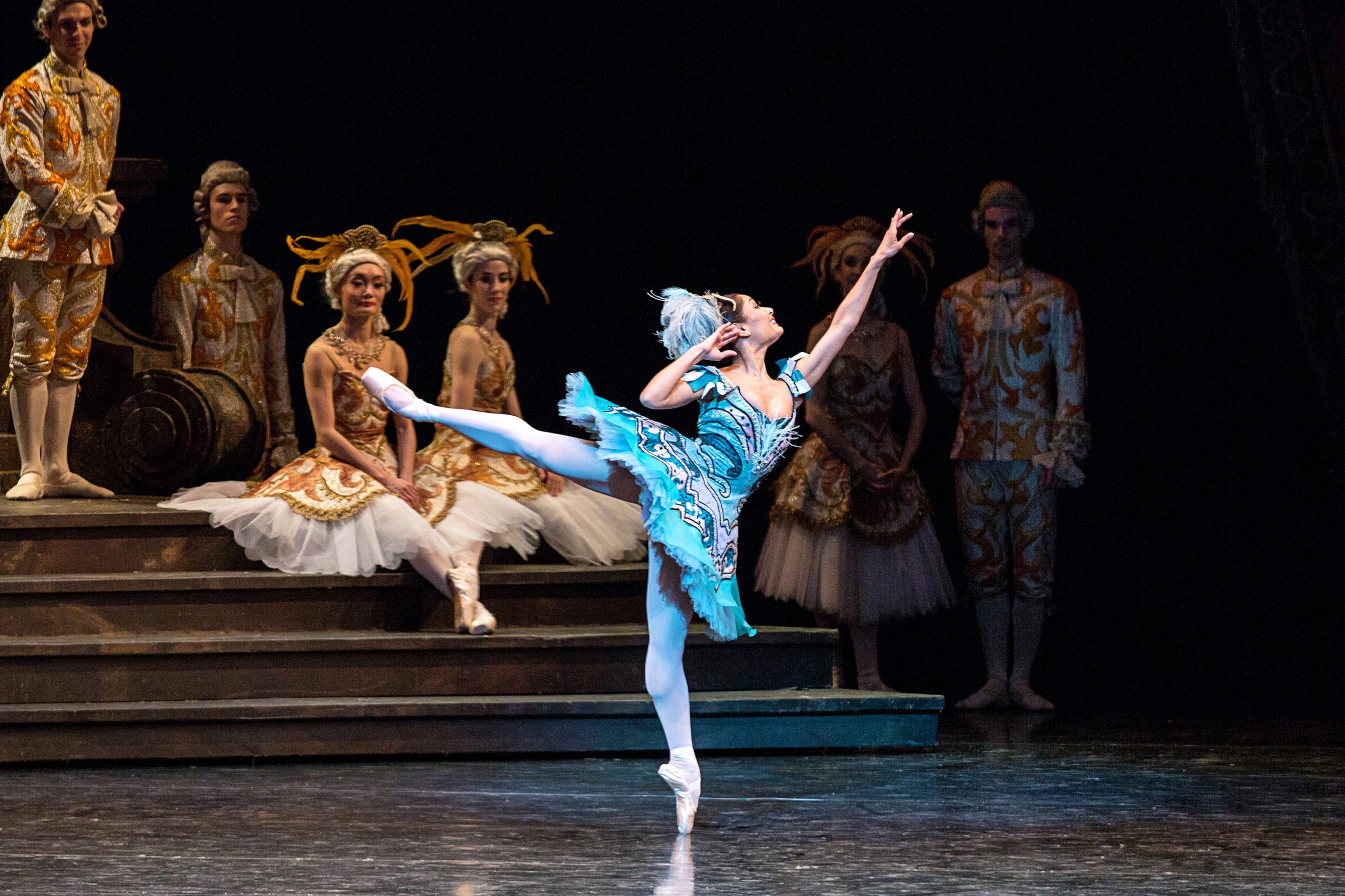 Tina Pereira performs in The Sleeping Beauty onstage. She wears a bright blue tutu and feathered headpiece, pink tights and pink pointe shoes. She performs a first arabesque with her right leg behind her, and she bends her right arm in so that her hand grazes her ear. Behind her, several dancers in grand costumes sit and stand on a staircase and watch her.