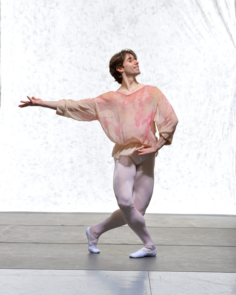 Christian Laverde Koenig wears a multit-colored, nuetral-toned loose-fitting, sheer long-sleeved top cinched at the waist. His tights are light pink and he wears white ballet slippers. He's shown in plié, with his right leg corssed over a slightly stretched left leg. His left hand is on his hip and his right arn is stretched out to the side with the palm facing up. He poses in front of a white backdrop.