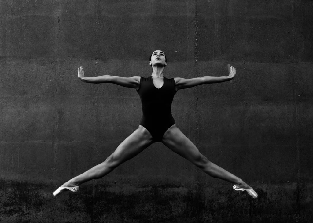 In this black and white photo, Sydnie Holmes wears a black leotard and jumps in an extra-wide second position, with her arms stretched wide to the side. She flexes her wrists and looks up toward the ceiling.
