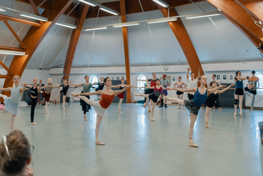 In a large ballet class in a big, airy studio, a group of female dancers practice first arabesque with their right leg up during class. They wear various dance outfits, tights and pointe shoes. In the back of the studio a group of dancers stand and watch.