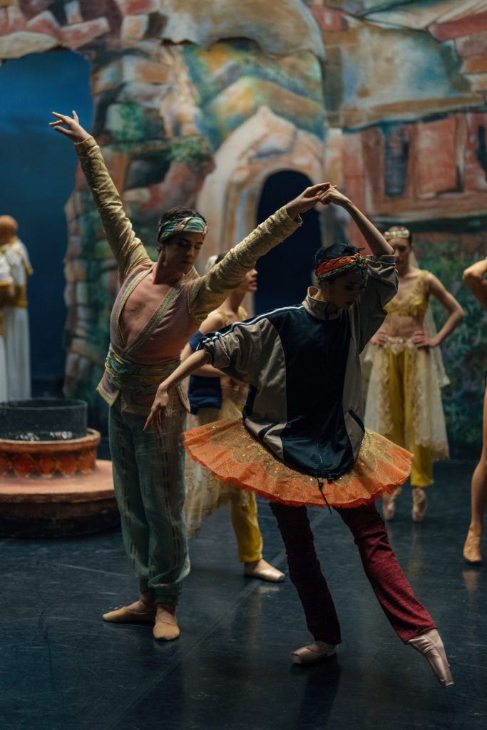 A male and female dancer practice backstage before a performance. The woman wears a tutu and pointe shoes and assorted warm ups, while the man wears blue harem pants, a gold tunic and blue and gold headpiece.