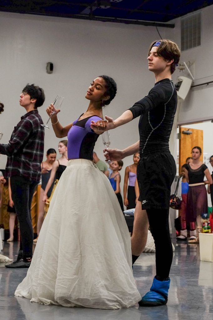 Tiana Ozolins, wearing a purple leotard and full, floor-length practice skirt, holds her male dance partner's left hand with her left hand and holds a champagne glass with her right during a crowded Nutcracker rehearsal. Her partner wears a black shirt and shorts and a legwarmer on his left leg.
