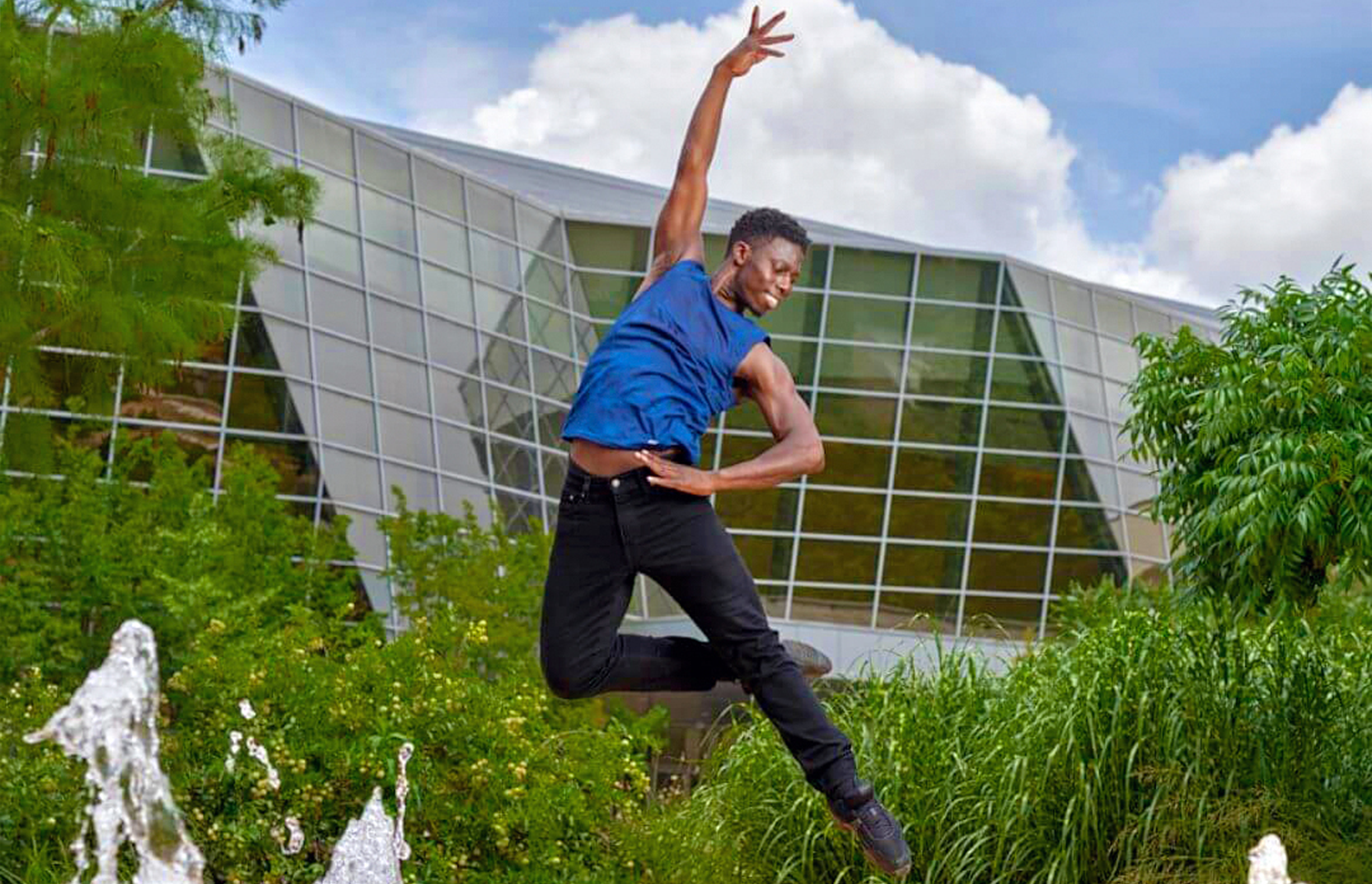 Brooklyn Mack soars in a retire saute, arms in high fourth, over a water fountain pad outside. A modern glass-window exterior building, trees and a blue sky are in the background. Mack wears a blue tank top, black tights and black ballet slippers.