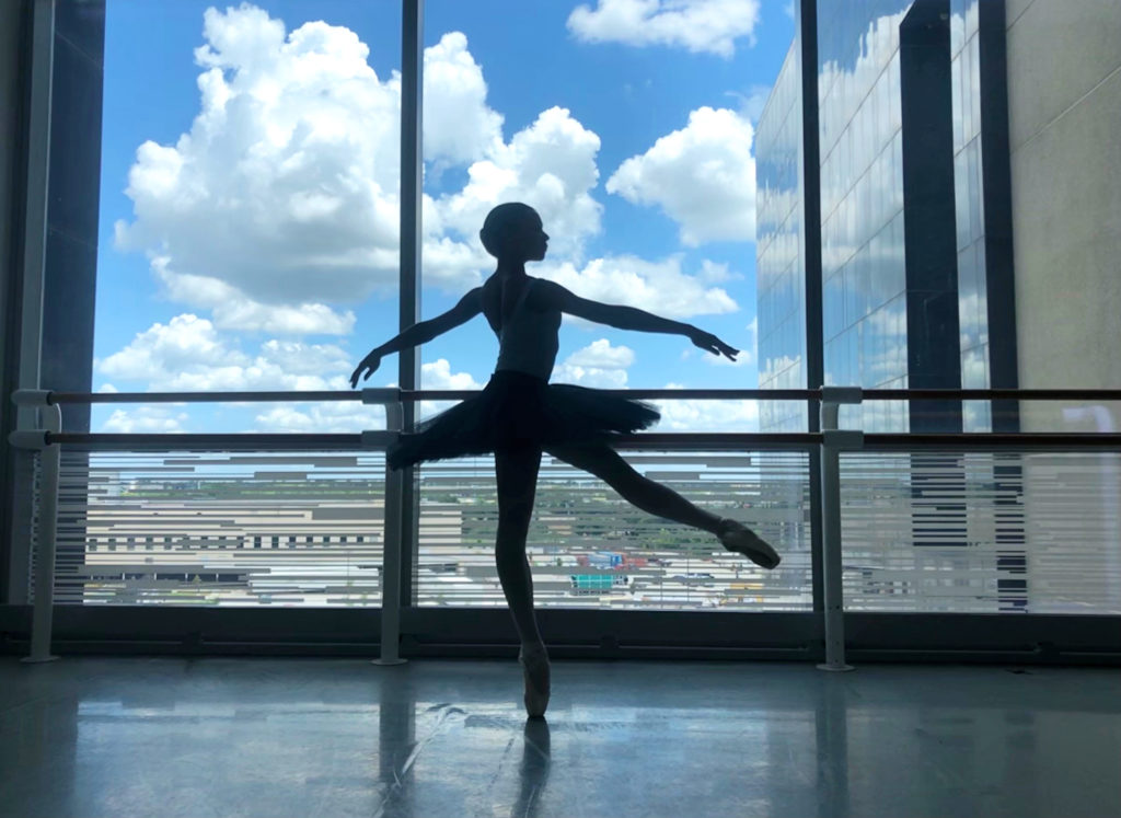 Gabrielle Miller is shown in silouette in front of a large floor-to-ceiling window in a ballet studio. She poses in profile facing her left on pointe, lifting her left leg in a degagé and stretching her right arm forward and left arm back. She wears a practice tutu and leotard.