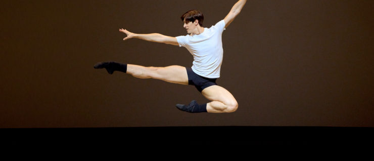 Xander Parish soars in a Russian pas de chat onstage. He wears a white fitted t-shirt, black dance shorts, black socks and black ballet shoes.