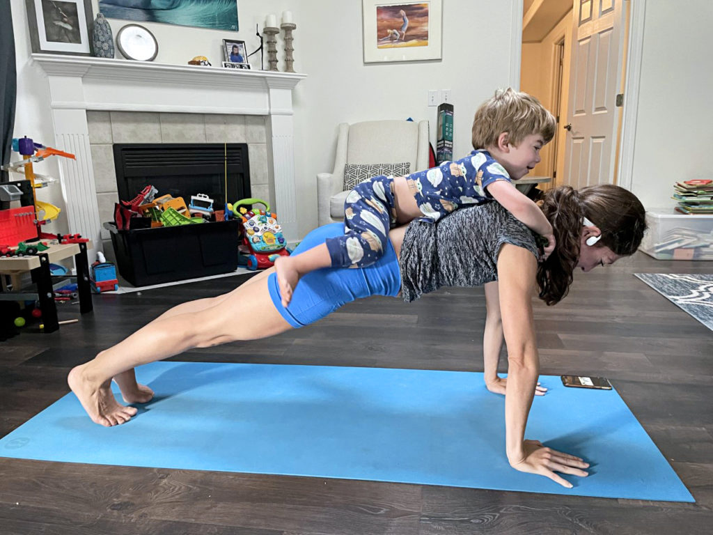 A woman holds a plank position while her toddler son lies on her back.