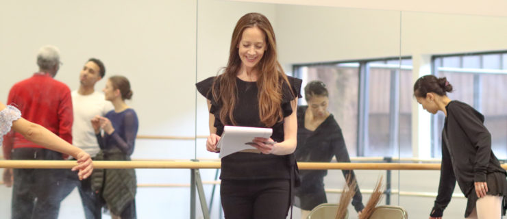 Maria Kowroski, wearing a black top and black pants, stands in the middle of a dance studio. She smiles and looks down at a pad of white paper. Behind her, dancers are reflected in the mirror, talking casually, while a female dancer walks toward the mirror and bends toward the floor.