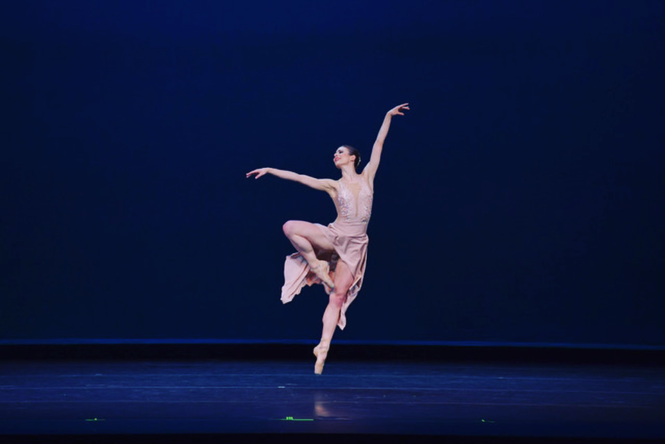 Ilse Kapteyn performs onstage in front of a dark blue backdrop. Her costume is a silky pink dress with a skirt that is higher in the front and longer in the back, and she wears pointe shoes. She performs a jump with her right leg in a retiré position and her left arm high and her right arm out to the side. She looks out over her right hand and smiles confidently.