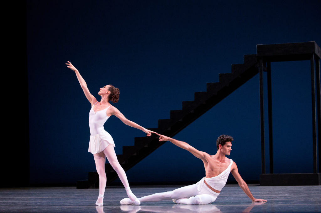 Tricia Albertson poses in tendu derriere facing stage right and touching her left pointer finger to Renan Cerdeiro's right pointer finger. Cerdeiro sits on the ground, on his left hip with his right leg extended and his body facing stage left. Behind them is a stark, black staircase. ALbertson wears a white leotard, short white skirt, pink tights and pointe shoes. Cerdeiro wears white tights and slippers and a white toga-style top.