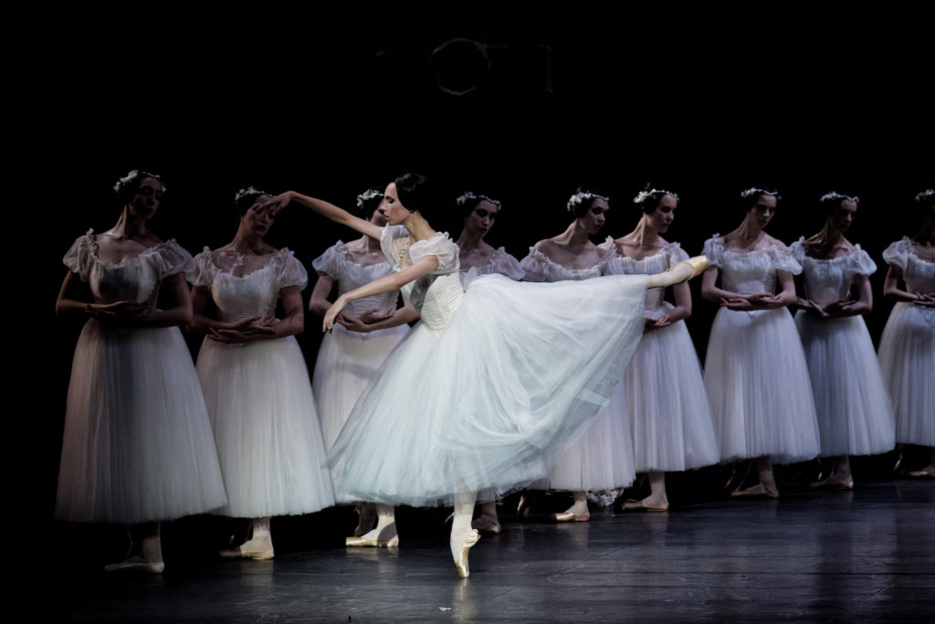 Bleuenn Battistoni, costumed in a white Romantic tutu with short puffed sleeves, performs a third arabeque on pointe towards stage right. A diagonal line of female corps dancers in the same costume stand in B plus behind her, crossing their wrists at the waist.