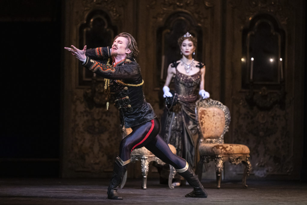 Vadim Muntagirov, dressed in rich costume, lunges forward on the diagonal with a distressed look on his face, eyes closed and left arm extended forward as if reaching for something. Behind him, an elegant woman in an elaborate evening gown stands and watches, hand rested on an ornate chair. They perform onstage amid other ornate scenic design.