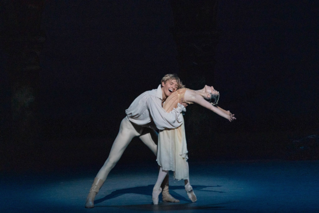 During a performance of Romeo and Juliet, Brooks Landegger holds Isadora Valero by the waist and presses his face against her body as she does a deep backbend while standing in fourth position on pointe. Landegger wears a white peasant blouse, white tights and tan boots, while Valero wears a calf-length, cream colored gown and beaded headpiece. Both dancers smile rapturously.