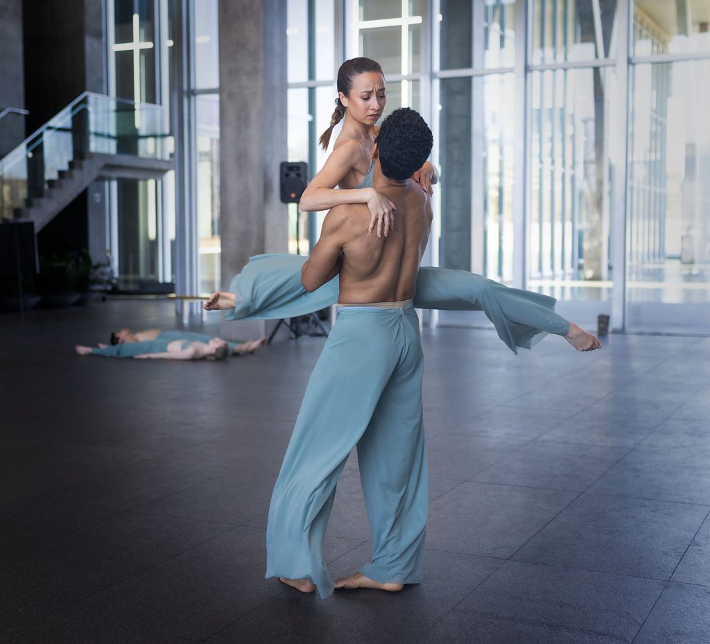 Amanda Fairweather and Joamanuel Velazquez wear flowy teal pants (Amanda also wears a teal bra top) as they dance together indoors in front of a wall of windows and a staircase. Joamanual lifts Amanda as she does a double attitude position, pressing down on his shoulders for support.