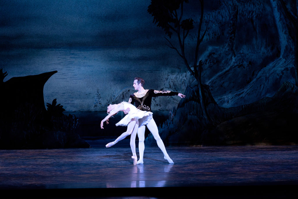 Alexandra Kochis and WIlliam Moore perform onstage in the ballet Swan Lake. Kochis poses on pointe, her right leg in a low attitude derriere, and does a deep back bend with her arms in a V shape while Moore holds her by the waist and looks over her. She wears a white tutu embellished with white feathers, pink tights and pink shoes. Moore wears a black and white velvet tuni, white tights and white ballet slippers, and holds his left arm out to the side. The backdrop shoes a wooded lake at nighttime.