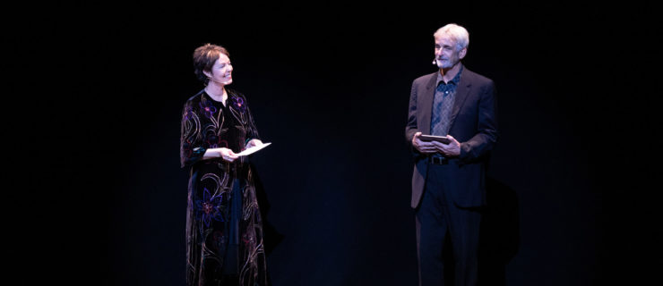 Susan Jaffe, on stage right, and Kevin McKenzie, to her left, stand onstage in front of a black curtain holding speech notes and with microphones attached to an earpiece on their right ears. Jaffe wears a long black velvet dress with a pattern of blue, purple and gold flowers. McKenzie wears a black sport coat and pants and gray buttong down shirt.