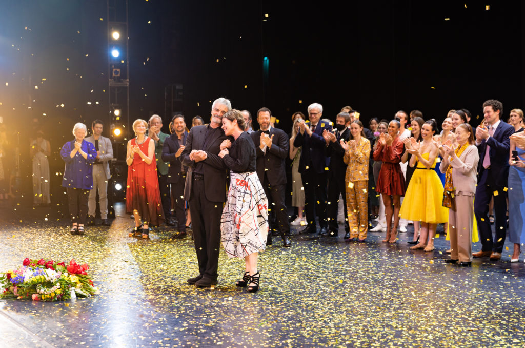 Kevin McKenzie and Susan Jaffe stand onstage, linking arms and huddling close as confetti rains down over them. Behind them, the dancers of American Ballet Theatre stand clapping and cheering, and a large heap of flowers rests on stage in front of the them. McKenzie wears a dark suit, while Jaffe wears a black top, calf-length white skirt with red and black patterns and strappy black high heels.