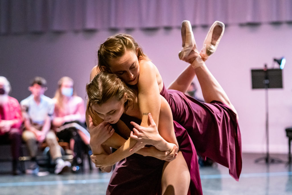 During a performance in the round, Nicole mcGinnis and Amber Neff, wearing burgundy gowns, perform a partnered move. McGinnis crouches and bends at the waist while Neff hugs her from behind and balances on her back, her legs raised and feet crossed. Audience members watch in the background.