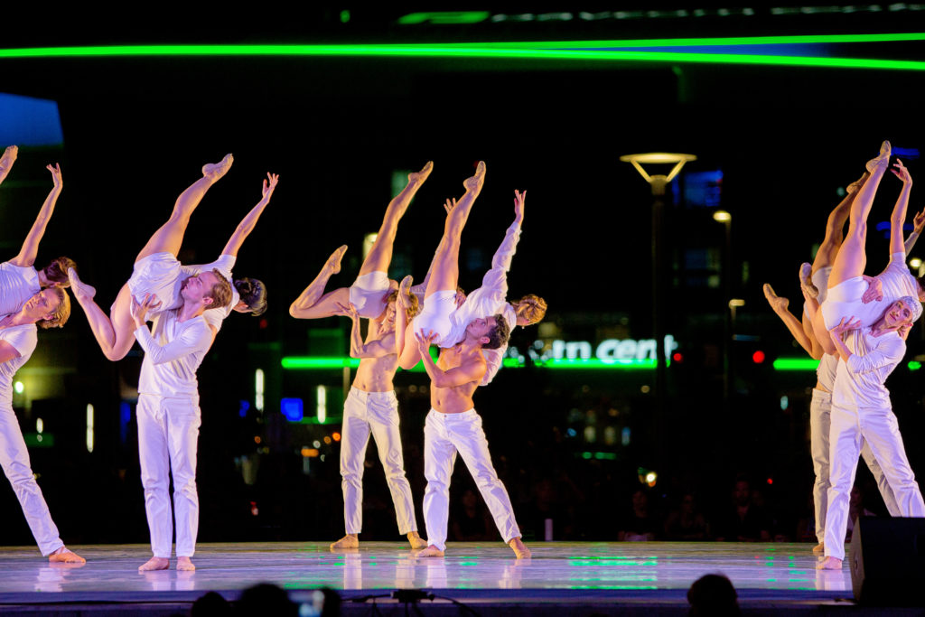 Six pairs of male and female dancers in tight white pants, shorts and tshirt costumes perform onstage at an outdoor theater. The men shift their weight to the right, lifting the women up to the side with their legs extended upward, facing away from the camera.