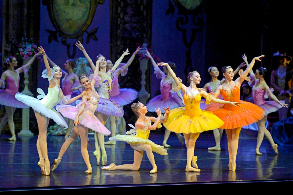 Six female dancers, a mix of professionals from State Ballet Theatre of Ukraine and local New Jersey students, pose onstage in the "Sleeping Beauty" Prologue fairy scene. They wear bright colored tutus in warm hues, pink tights and pointe shoes, fairy wings and tiaras. Other dancers in pink tutus pose behind them in the background.