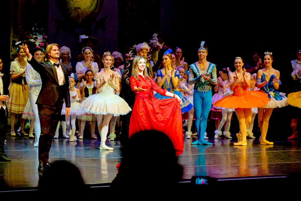 Artem Yachmennikoff, Ekaterina Vaganova-Yachmennikova, members of State Ballet Theatre of Ukraine and local ballet students clap onstage during bows after the SBToU performance of "Sleeping Beauty." Artem wears a black tuxedo, Ekaterina wears a long red dress, and the dancers are in costume as the audience applauds in the foreground.