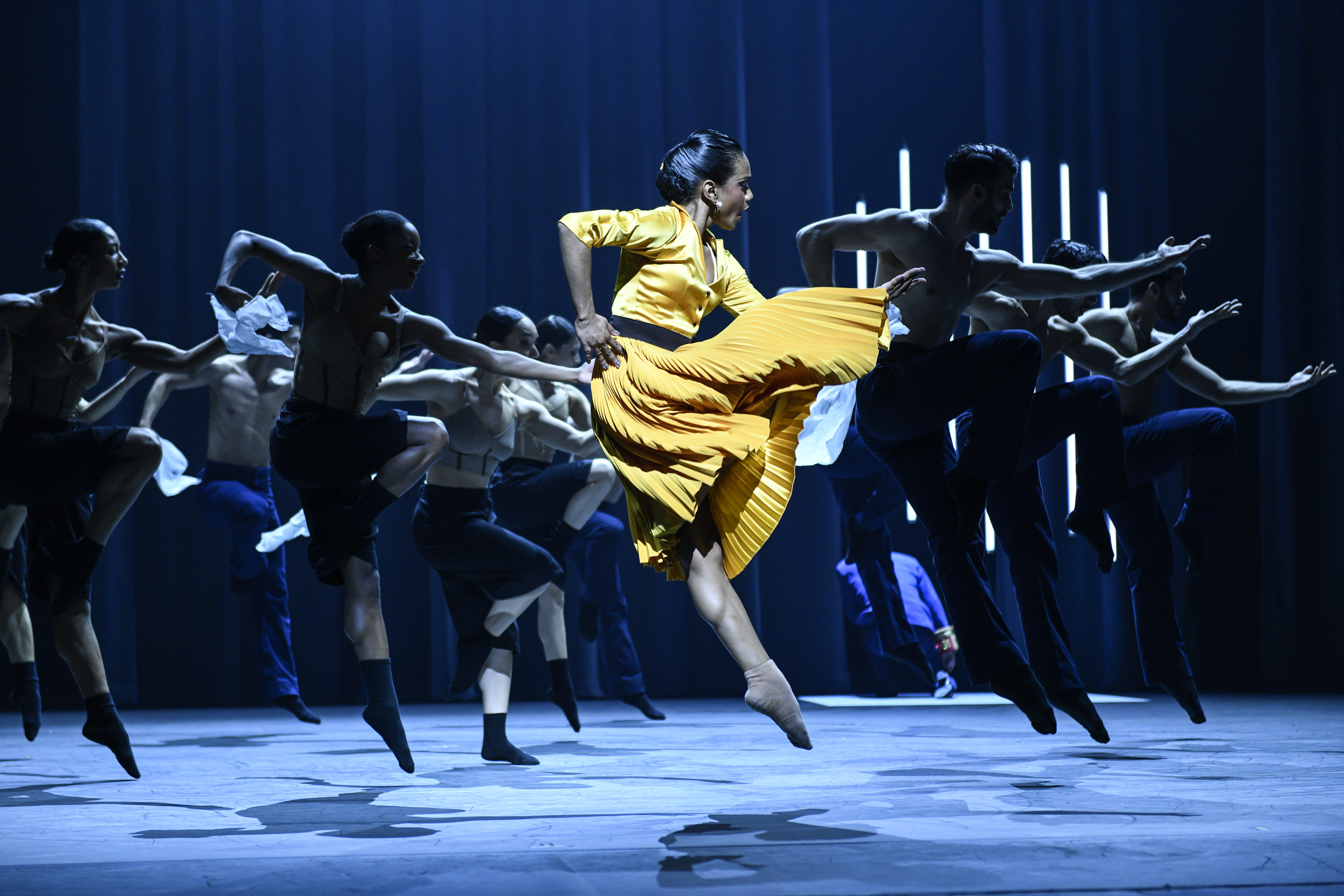 Dandara Vega, costumed in a bright yellow dress with leated skirt, jumps in a parallel passé. She is spotlit and dances at the front of a large group, who do the same step behind her.