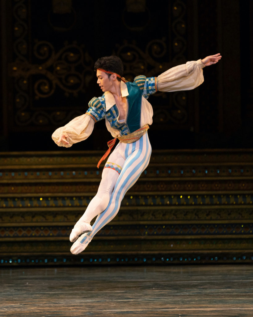 During a performance, Elwince Magbitang performs a brisé to his right. He wears a billowy off-white shirt with blue-striped trim, a thin orange headband, white tights with blue-stipes along the left leg and white ballet slippers. A glittering staircase is upstage of him in the background.