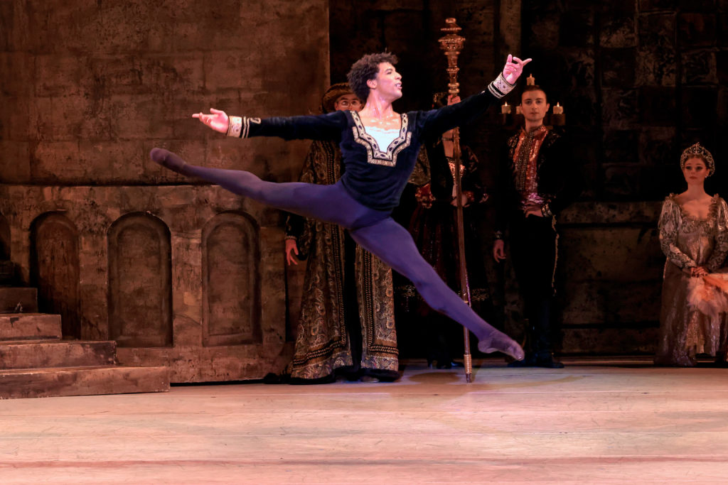 Gian Carlo Perez performs onstage during a grand ballroom scene from Swan Lake. He wears a dark tunic, dark tights and ballet slippers and performs a high sissonne fermee en avant towards stage left. Three dancers in medieval costumes sit and watch in the background.