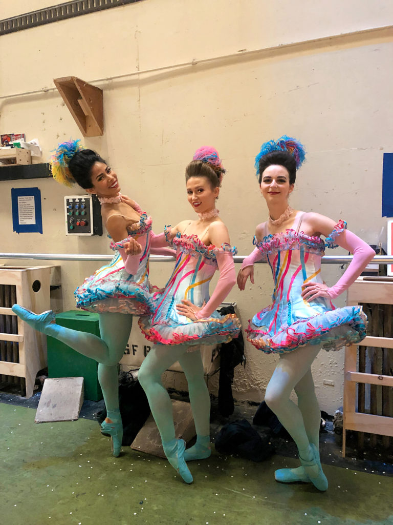 Gabriela Ginzalez and two other female dancers pose for a casual photo while dressed in colorful tutus. The dancers wear blue tights and pointe shoes and colorful, feathered headpieces.