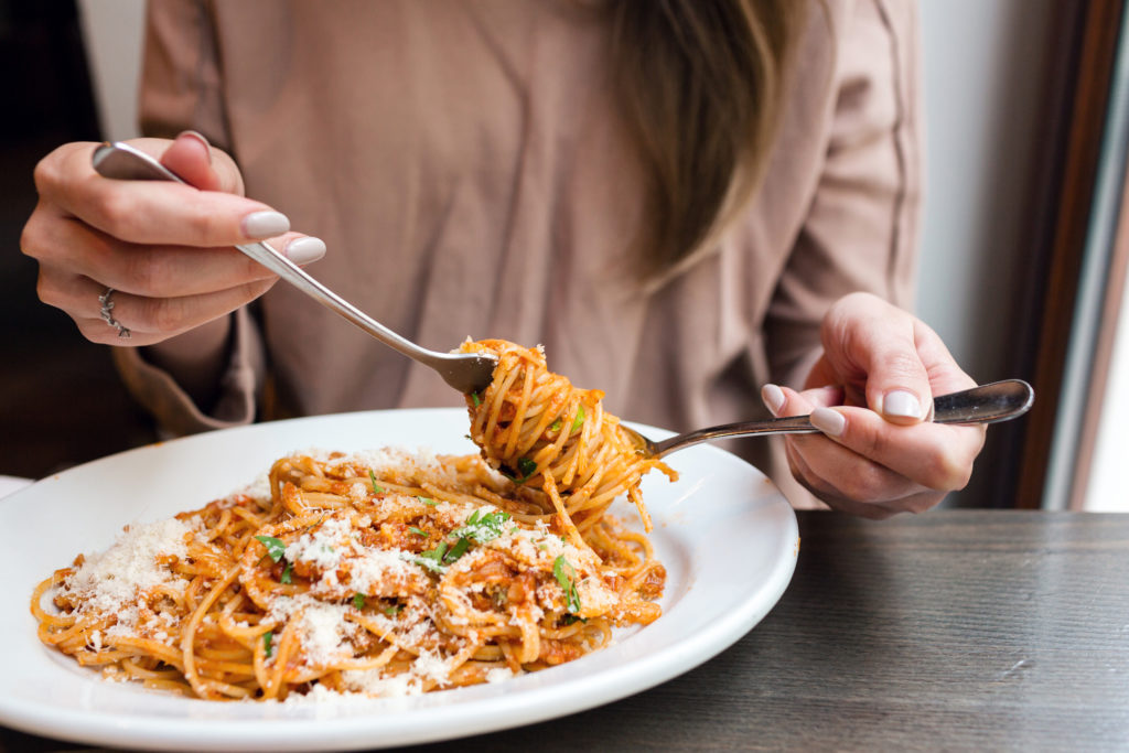 A woman eats Italian pasta with tomato, meat sauce and Parmesan cheese. She winds it around a fork with a spoon.
