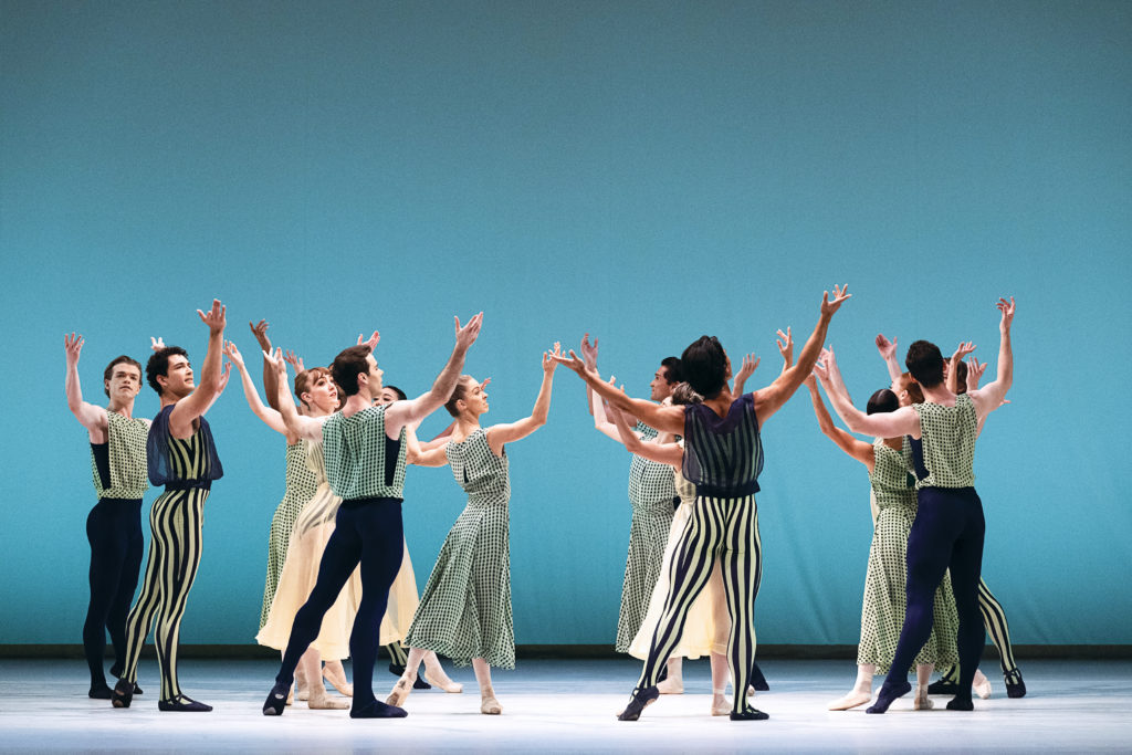 A group of male and female dancers form a circle onstage, standing in tendu derrierre with their arms lifted up. They wear variious costumes in shades of green, cream and black.