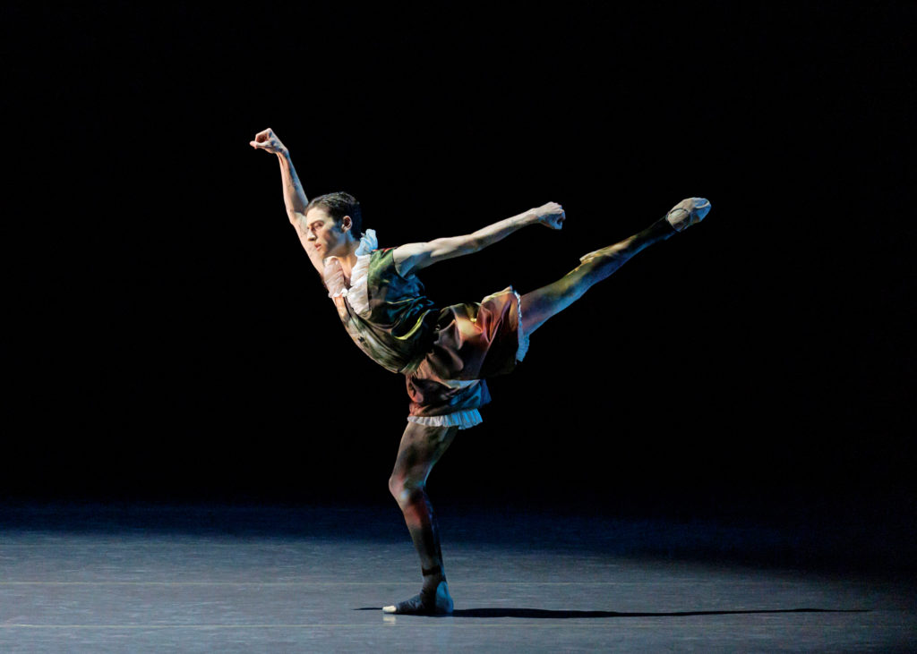 Jonathan Fahoury, dressed in a dark-toned, multi-colored tights and costume with a white ruffled collar and white ruffled shorts, does a first arabesque allongé with his left leg up during a performance. He clenches his fists and looks down toward the ground. Behind him, the stage is dark.