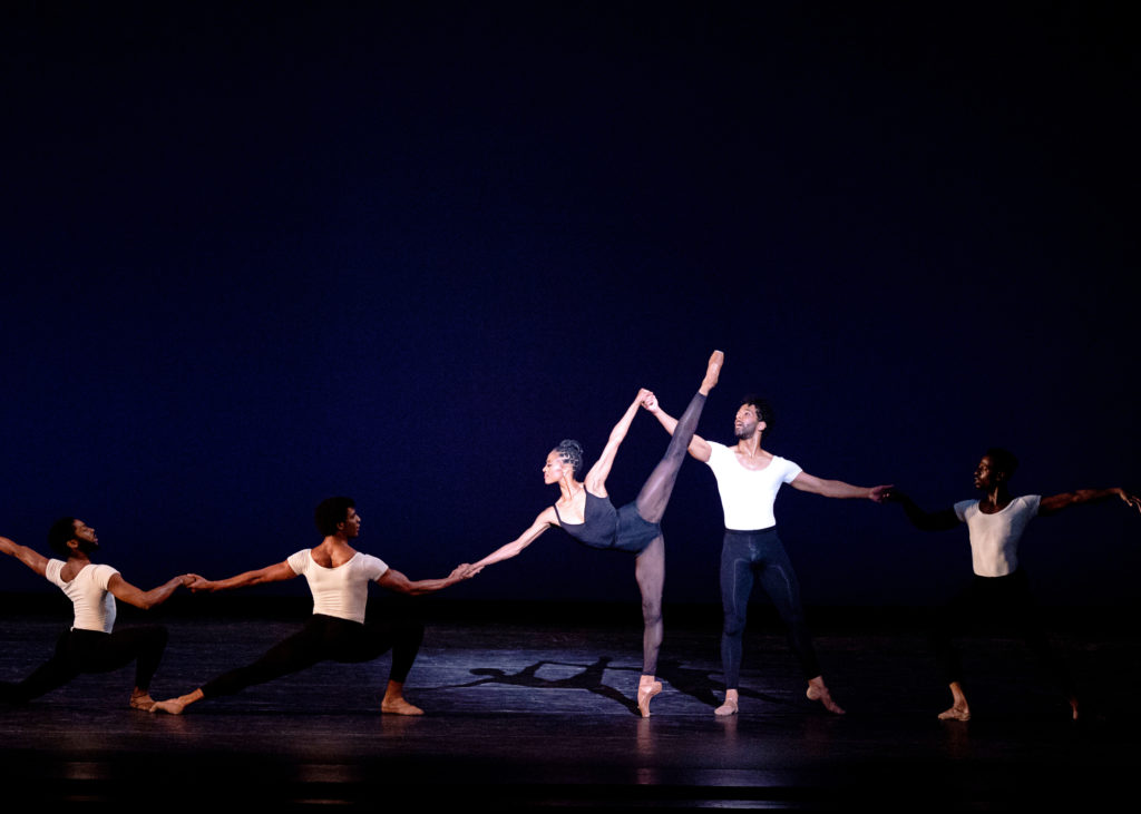 Katlyn Addison, wearing a black leotard, black tights and brown pointe shoes, performs a penché arabesque on pointe with her left leg raised, while two male dancers hold each of her hands so she is supported. Two men lunge deeply in front of her, holding hands, while the other man holding her hand stands behind her in tendu and links hands with another male dancer lunging in fourth position. The men wear white T-shirts, black tights and tan ballet slippers. The stage is dark, with Addison spotlit.