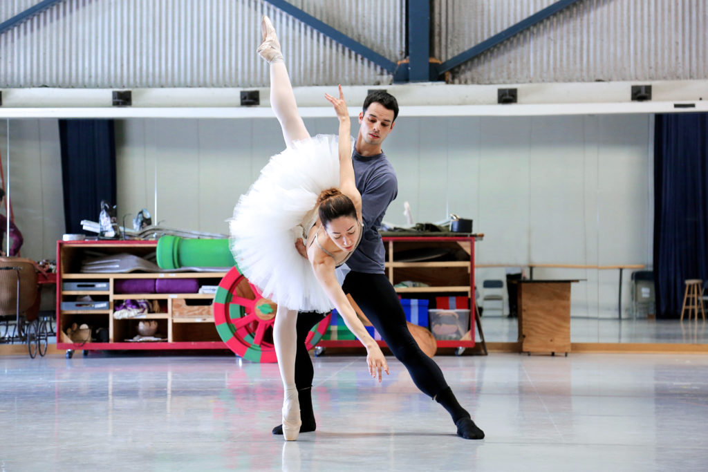Kristina Kadashevych and Aleksey Babayev rehearse a pas de deux in a bright studio with props in the background. Kadashevych wears a leotard and white practice tutu with pink ballet tights and pointe shoes, and she poses in an arabesque penche toward the audience. Babeyev supports her and wears a blue shirt with black ballet tights and flats.