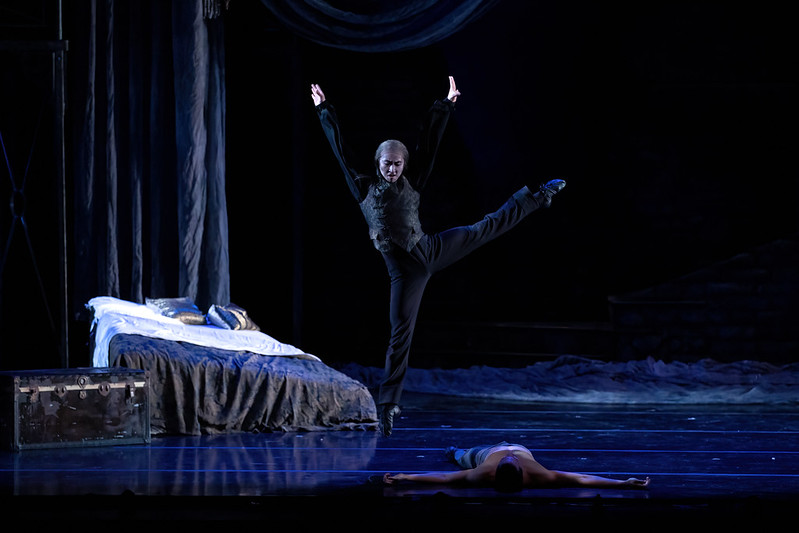 Liang Fu, dressed as Dracula in dark, gothic clothing, does a wide a la seconde sauté onstage, looking down at Lamin Pereira, who lies prone on the floor. There is a bed and old trunk behind them onstage.