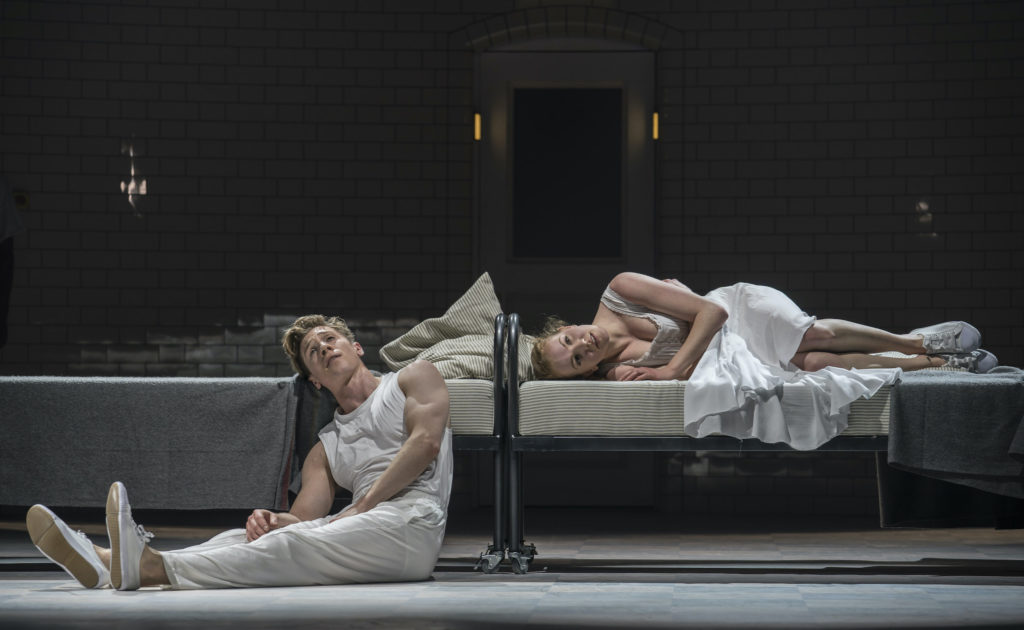 Dancers Andrew Monaghan and Seren Williams are shown onstage during a performance set in an a mental institution. Managhan, dressed in a white tank top, pants and sneakers, sits on the ground and rests his head against a spare twin bed, looking wistfully towards the sky. Williams lays on another twin bed next to his, curling up her legs, and wearing a white dress and sneakers.