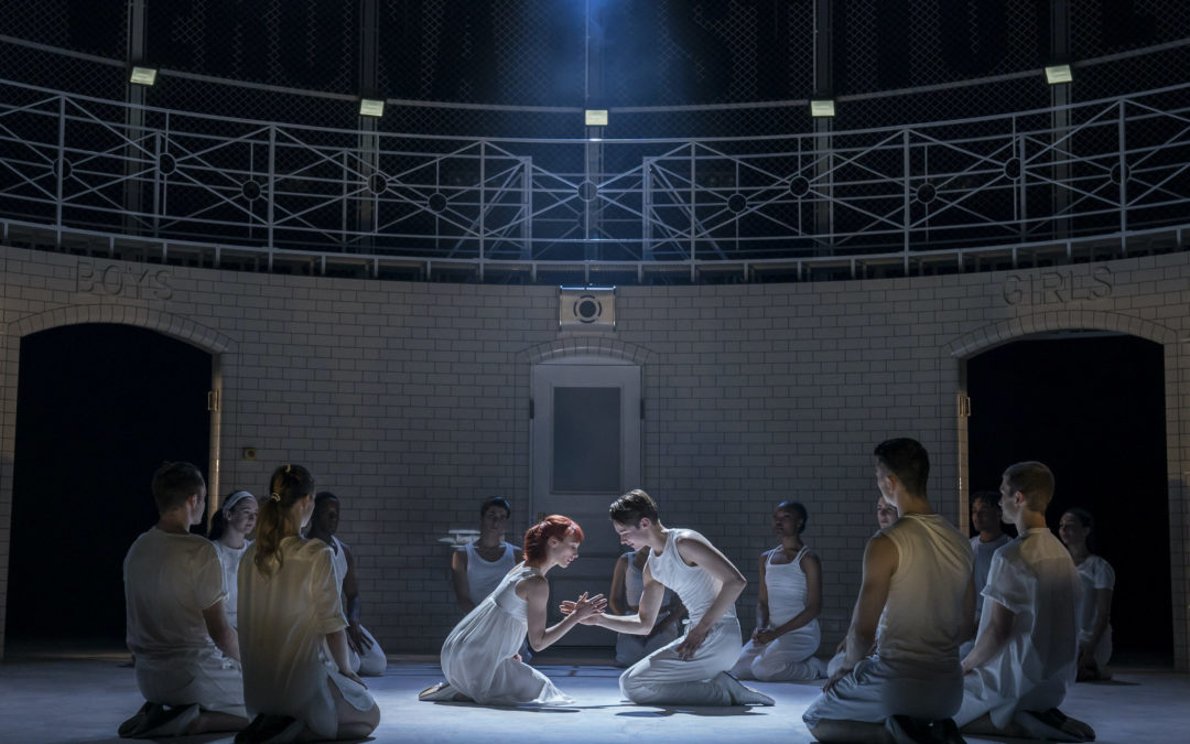 Cordelia Braithwaite and Paris Fitzpatrick, dressed all in white, kneel center stage during a performance, facing each other and joining right hands. A spotlight shines on them from above, and a circle of other dancers, also dressed in white, kneels on the ground surrounding them.