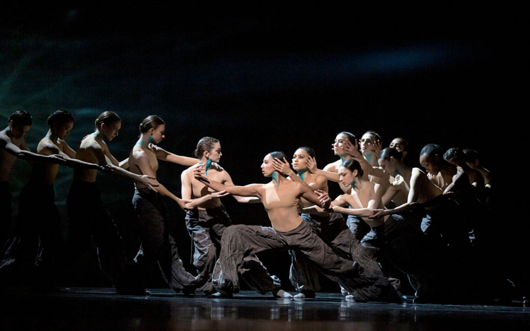 Amanda Morgan does a deep lunge in prfile, facing stage right, while a large group of female dancers form a chain on either side of her, holding arms. They all wear nude leotards, baggy dark pants, and their throats are painted blue. The stage is dark behind them.