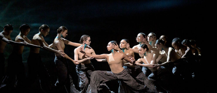 Amanda Morgan does a deep lunge in prfile, facing stage right, while a large group of female dancers form a chain on either side of her, holding arms. They all wear nude leotards, baggy dark pants, and their throats are painted blue. The stage is dark behind them.