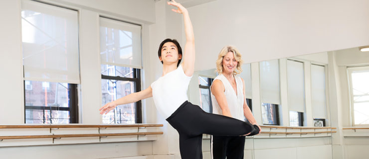 In a sunny studio, Asahi Hagihara poses in an attitude derriere as Stacey Calvert stands behind him and holds raised his left leg. Hagihara wears a white T-shirt, black tights and black ballet slippers, while Calvert wears dark exercise pants, black shoes and light, V-neck shirt.